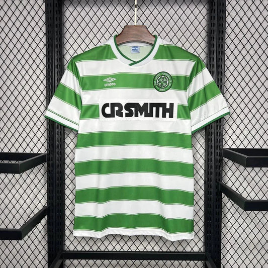 Front view of a classic Celtic FC football shirt with green and white hoops, Umbro logo, CR Smith sponsor, and club crest, hanging on a wooden hanger against a mesh background.