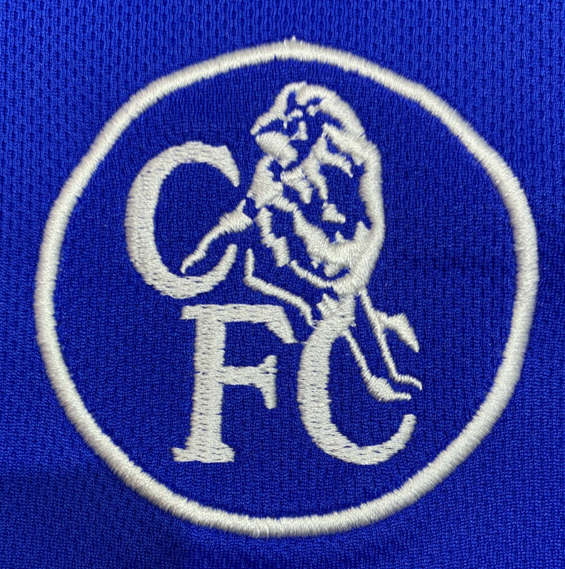 Close-up of the retro Chelsea FC circular club crest featuring a lion and the letters CFC, embroidered in white on blue fabric.