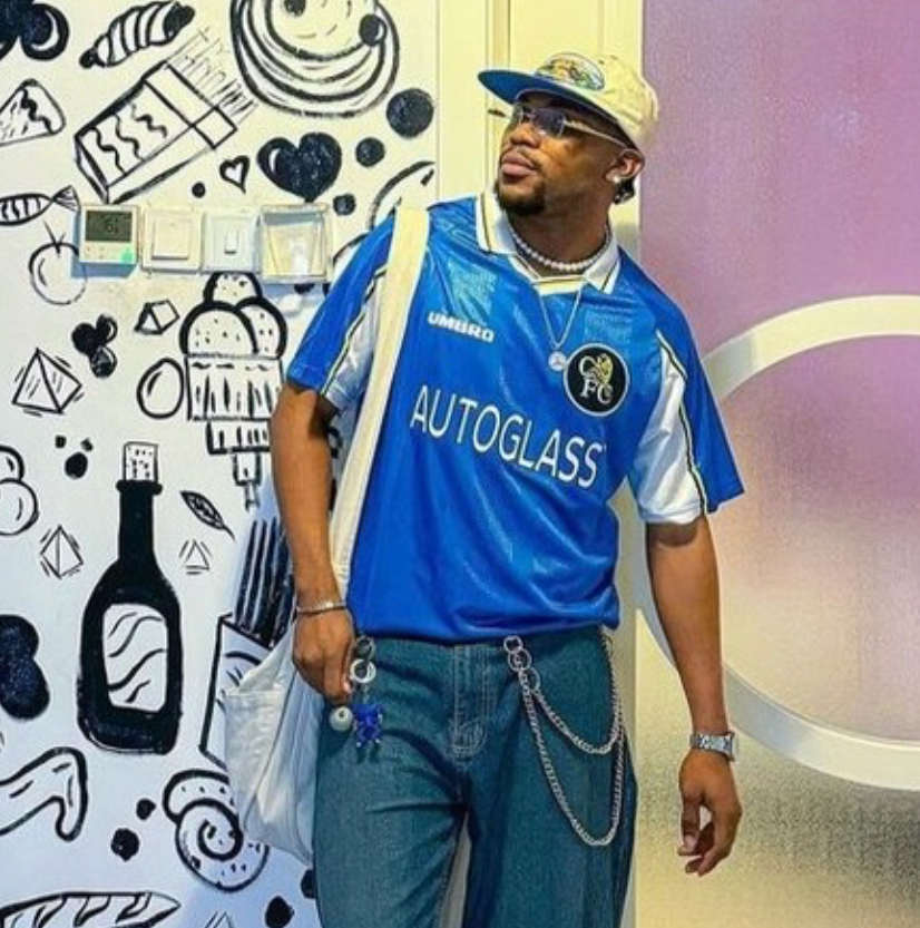 A person wearing a vintage blue Chelsea football jersey with "Autoglass" sponsor, wide-leg jeans, black shoes, and accessories including a chain, watch, necklace, and a white bag, standing against a wall with black-and-white food-themed illustrations and patterned floor tiles.