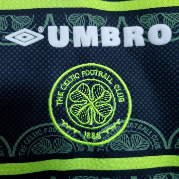 Close-up of a 1999 Celtic FC football shirt featuring the Umbro logo, club crest, and neon yellow horizontal stripes on a dark background.