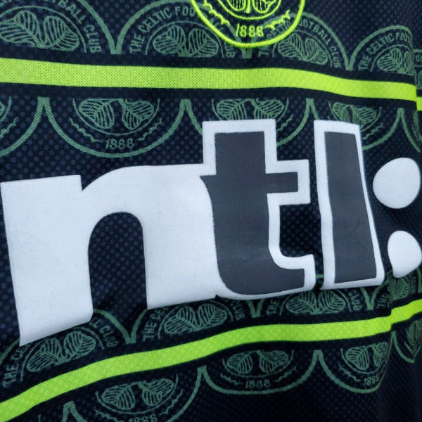Close up of the 1999 Celtic FC football shirt featuring the Umbro logo, club crest, and neon yellow horizontal stripes on a dark background.