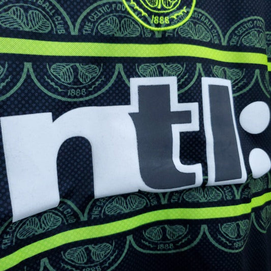 Close up of the 1999 Celtic FC football shirt featuring the Umbro logo, club crest, and neon yellow horizontal stripes on a dark background.